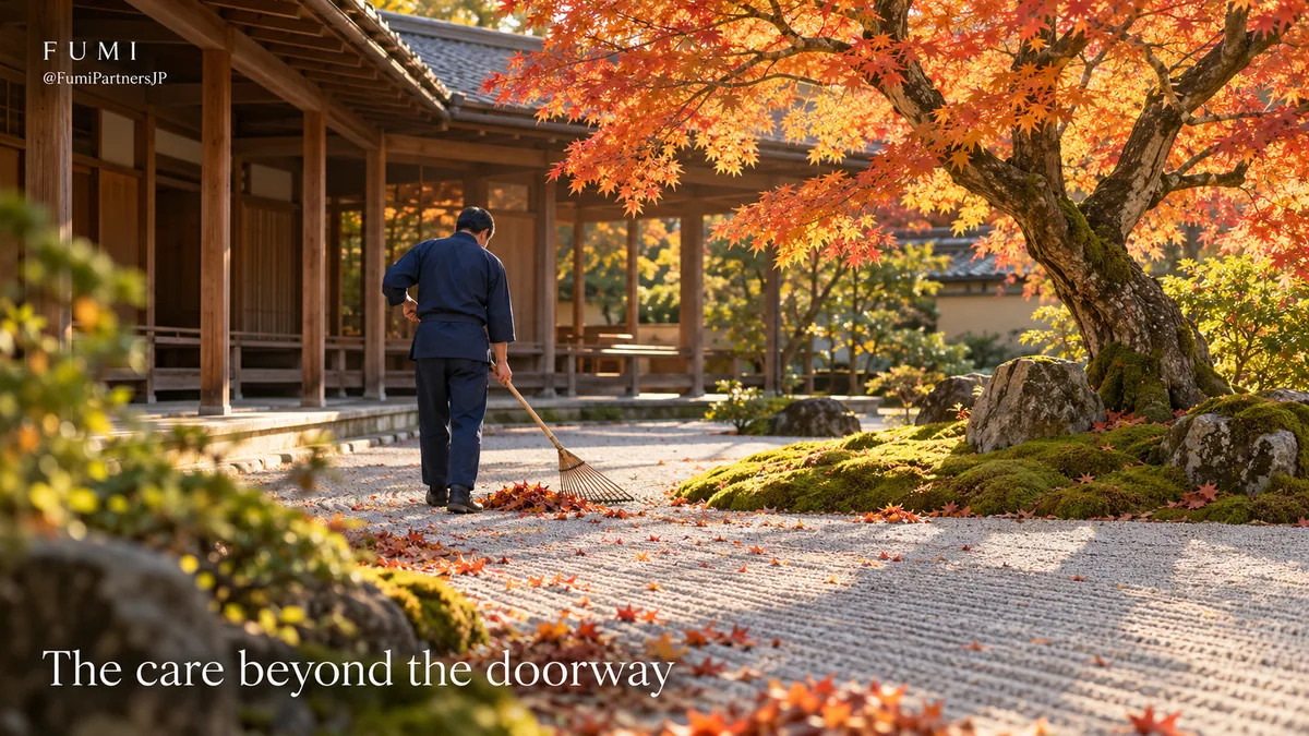 A gardener in plain indigo workwear rakes fallen leaves in a sunlit Japanese courtyard beneath an orange maple.